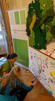 Two women working on a map of Czechia in front of the Heinrich Böll Foundation poster. On top: Outline of Czechia filled with (artificial) trees symbolizing forests in Czechia.